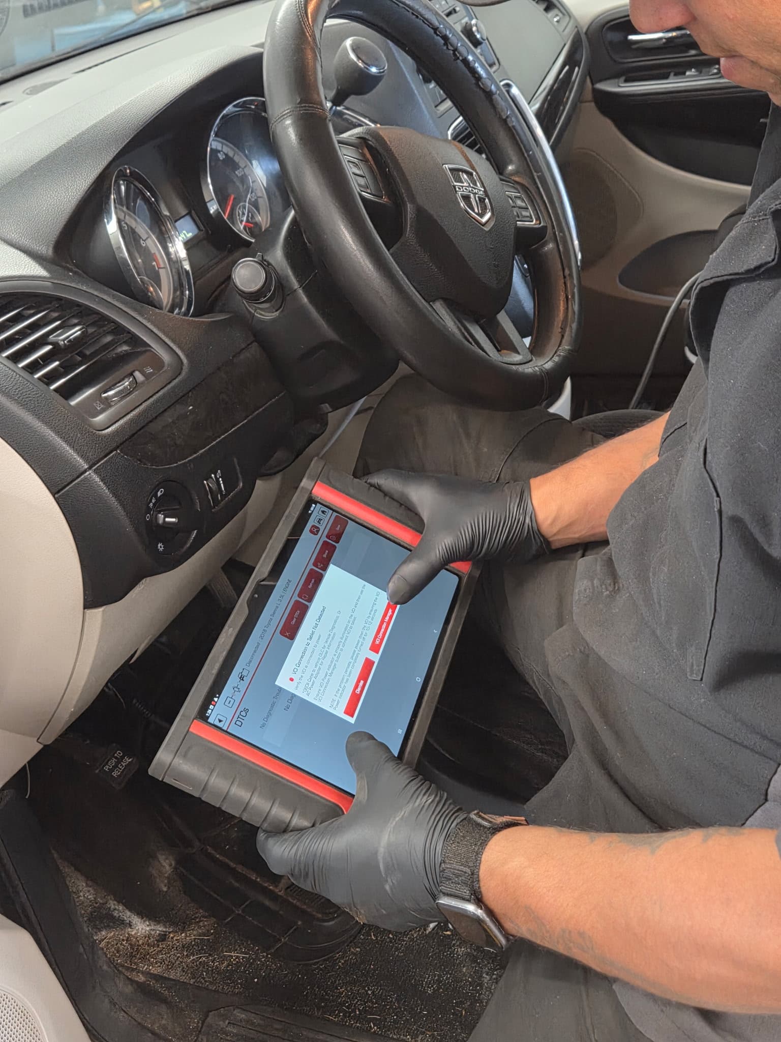 Mechanic in black gloves uses a diagnostic tablet tool inside a car's driver seat.