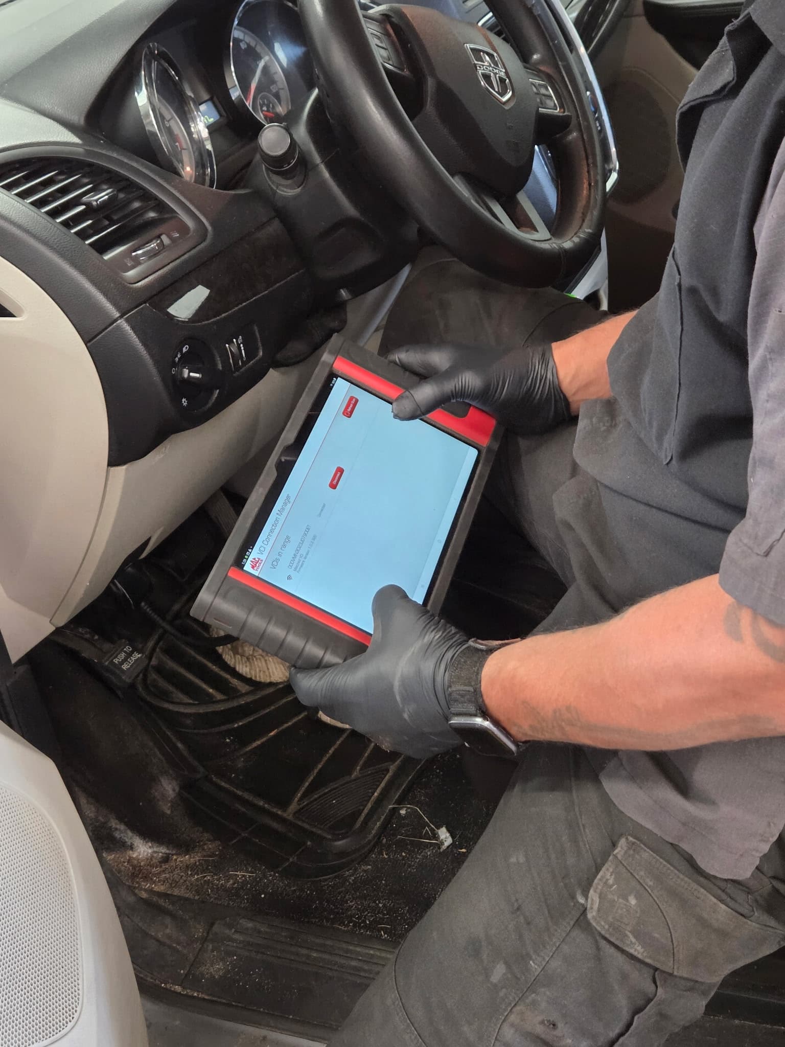 Mechanic in black gloves uses a diagnostic tablet inside a Dodge vehicle's driver seat.