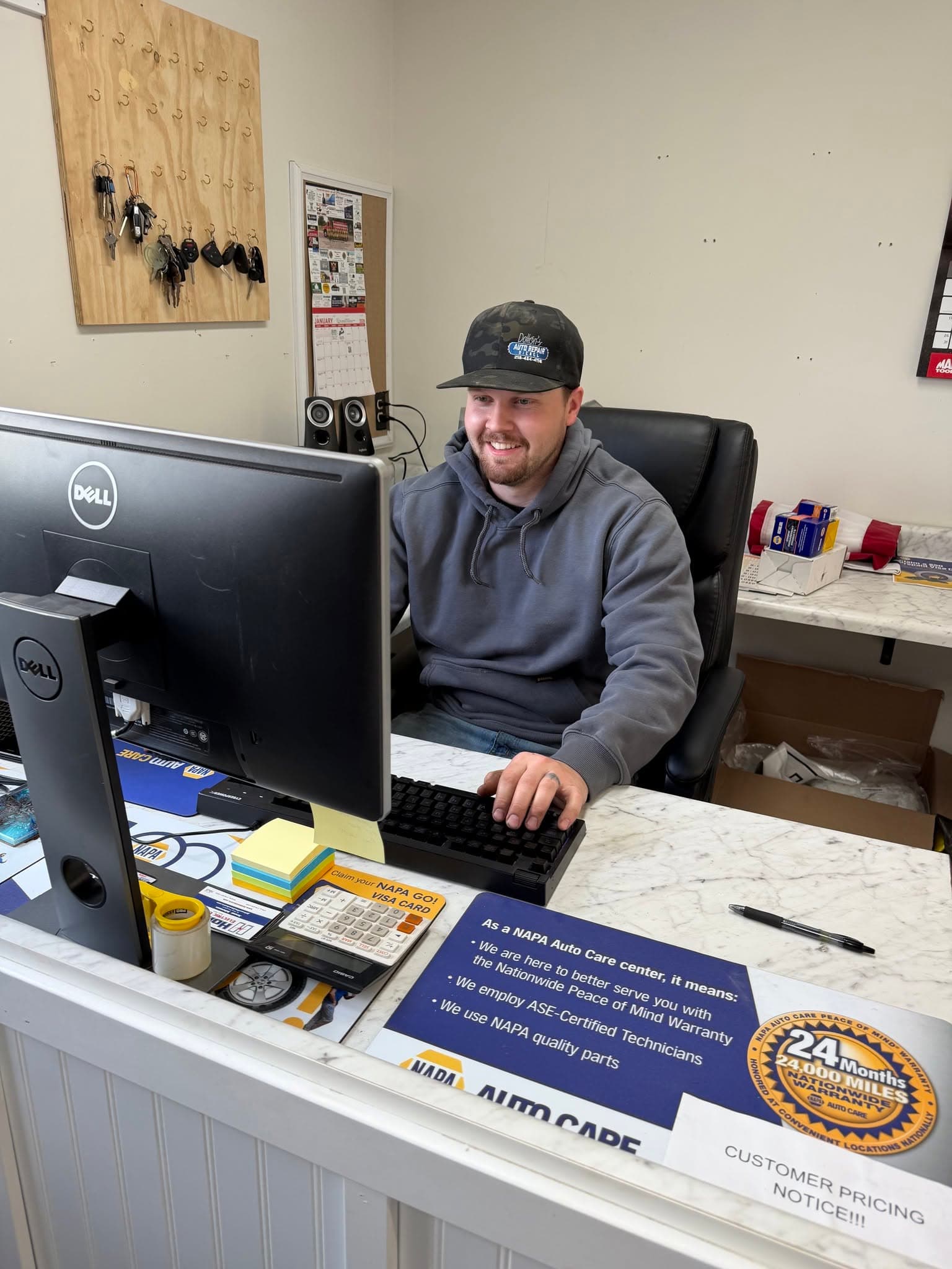 Smiling man in a cap and hoodie using a computer at an auto repair desk.